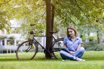 Mujer joven sentada cruzando el césped junto a la bicicleta utilizando tabletas digitales - foto de stock
