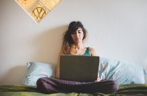 Vorderansicht einer erwachsenen Frau, die mit einem Laptop im Schneidersitz auf dem Bett sitzt — Stockfoto