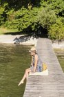 Vista lateral da jovem mulher sentada no cais de madeira usando chapéu de panamá olhando para longe, Schondorf, Ammersee, Baviera, Alemanha — Fotografia de Stock