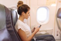 Mujer joven en el avión eligiendo la música en el teléfono inteligente - foto de stock