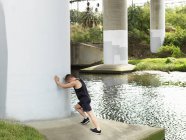 Young man exercising beside river, leaning against column, stretching — Stock Photo