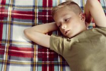 Vue grand angle du garçon allongé sur une couverture de pique-nique mains derrière la tête regardant loin — Photo de stock