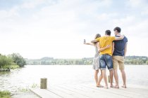 Three young adults standing on jetty, taking self portrait, using smartphone, rear view — Stock Photo