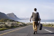Rear view of young man with guitar case walking on coastal road — Stock Photo
