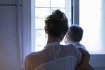 Rückansicht der erwachsenen Frau und der kleinen Tochter, die aus dem Wohnzimmerfenster schauen — Stockfoto