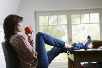 Зріла жінка з ногами на столі, п'є каву — стокове фото