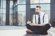 Stylish businessman sitting cross legged making diary notes from smartphone outside office — Stock Photo