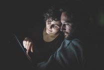 Пара сидящих в темной комнате, глядящих на цифровой планшет — стоковое фото