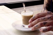 Mão tatuada de jovem e copo de café na mesa de café — Fotografia de Stock