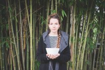 Вид спереду молода жінка стоїть перед бамбуковим гаєм, тримаючи керамічну тарілку, дивлячись на камеру — стокове фото