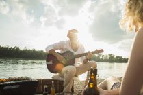 Young man sitting by lake playing guitar — Stock Photo