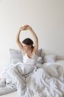 Зріла жінка в жилеті сидить у ліжку під ковдрою руки підняті розтягування — стокове фото
