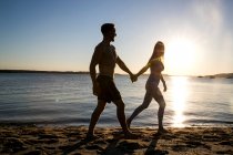 Зворотній бік молода людина і дівчина, що йде рукою на пляжі під час заходу сонця — стокове фото