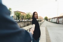 Vista sobre el hombro de una mujer joven sosteniendo la mano del novio por el canal de la ciudad - foto de stock