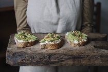 Woman holding chopping board, with ricotta, avocado and walnut bruschetta on top, mid section — Stock Photo