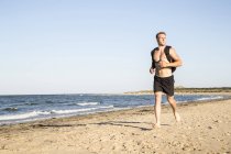 Joven corredor masculino en pantalones cortos y mochila corriendo por la playa - foto de stock