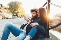 Hipster couple sitting looking at smartphone by city canal — Stock Photo
