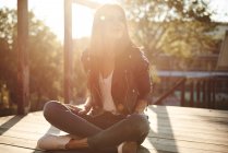 Jeune femme assise jambes croisées sur une terrasse en bois, regardant ailleurs, souriant — Photo de stock