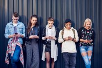 Reihe von fünf jungen erwachsenen Freunden lehnt an schwarzer Wand und schaut auf Smartphones — Stockfoto