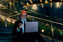 Mature businessman sitting on steps with laptop and smartphone — Stock Photo