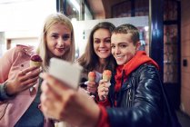 Três jovens mulheres com cones de sorvete levando selfie smartphone na rua da cidade — Fotografia de Stock