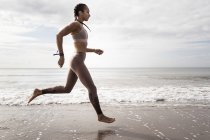 Боковой вид молодой бегуньи, бегущей босиком вдоль берега воды на пляже — стоковое фото
