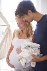 Embarazada pareja sosteniendo pila de ropa de bebé en vivero - foto de stock