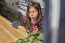 Молодая девушка сидит за столом, пьет сок из бутылки с помощью соломы — стоковое фото