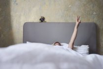 Woman in bed, hiding under covers, arm in air, hand showing peace sign — Stock Photo