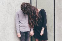 Young couple outdoors, standing against wall, holding hands, hair covering their faces, — Stock Photo
