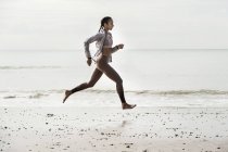 Vista lateral de una joven corredora corriendo descalza a lo largo del borde del agua en la playa - foto de stock