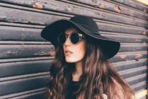 Портрет молодої жінки, що стоїть поруч із затвором, з капелюхом і сонцезахисними окулярами — стокове фото
