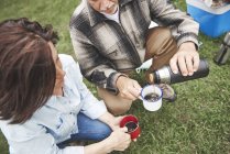 Mature couple on grass pouring hot drink from drinks flask — Stock Photo