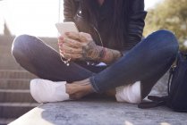Young woman sitting on wall, using smartphone, tattoos on hand, low section — Stock Photo