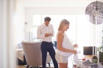 Homme regardant smartphone et petite amie enceinte pliant blanchisserie dans le salon — Photo de stock