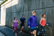 Four young adult runners running along city sidewalk — Stock Photo