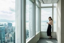 Business woman in skyscraper office window using smartphone — Stock Photo