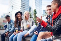 Cinco jovens amigos adultos sentados na parede conversando na cidade — Fotografia de Stock