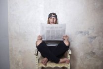 Portrait of woman sitting cross-legged on chair, reading newspaper — Stock Photo