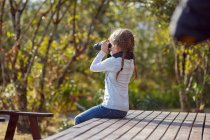 Girl in rural setting, sitting on decking, looking through binoculars — Stock Photo