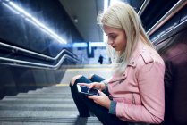 Молода жінка сидить на підземних сходах, дивлячись на смартфон — стокове фото