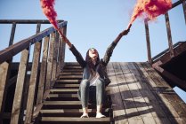 Young woman sitting on wooden steps, holding hand flares, red smoke pouring from flares, low angle view — Stock Photo