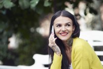 Молода жінка сидить на відкритому повітрі, використовує смартфон, посміхається, татуювання на руці та шиї — стокове фото