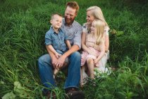 Семья, сидящая в высокой траве вместе улыбаясь — стоковое фото