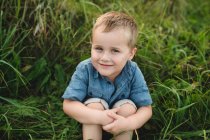 Retrato de menino sorridente sentado na grama alta olhando para a câmera — Fotografia de Stock