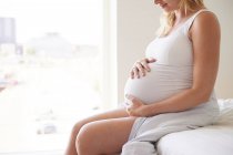Беременная женщина сидит на кровати с руками на животе — стоковое фото