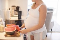 Coupé coup de jeune femme enceinte tranchant pastèque dans la cuisine — Photo de stock
