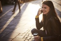 Jeune femme assise à l'extérieur, tenant une tasse de café, utilisant un smartphone, tatouages sur les mains — Photo de stock