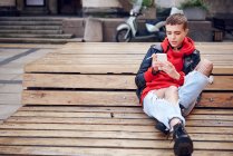 Jovem legal com cabelo curto olhando para smartphone e fumando cigarro no banco da cidade — Fotografia de Stock