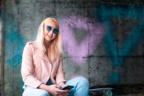 Portrait of young blond female skateboarder wearing sunglasses at skateboard park — Stock Photo
